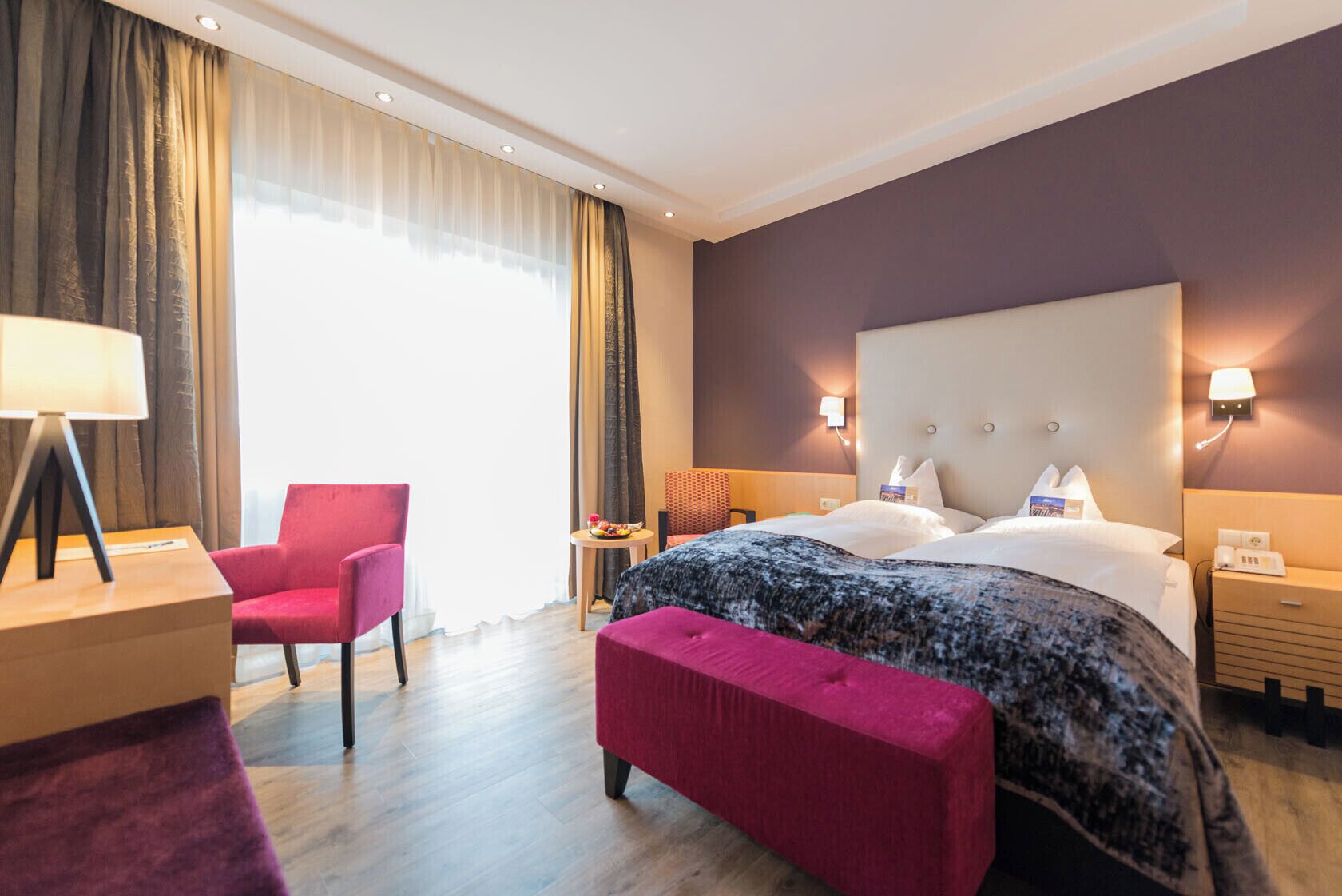 Une chambre d’hôtel avec un grand lit, un fauteuil rouge, une petite table avec des fruits et une lampe.