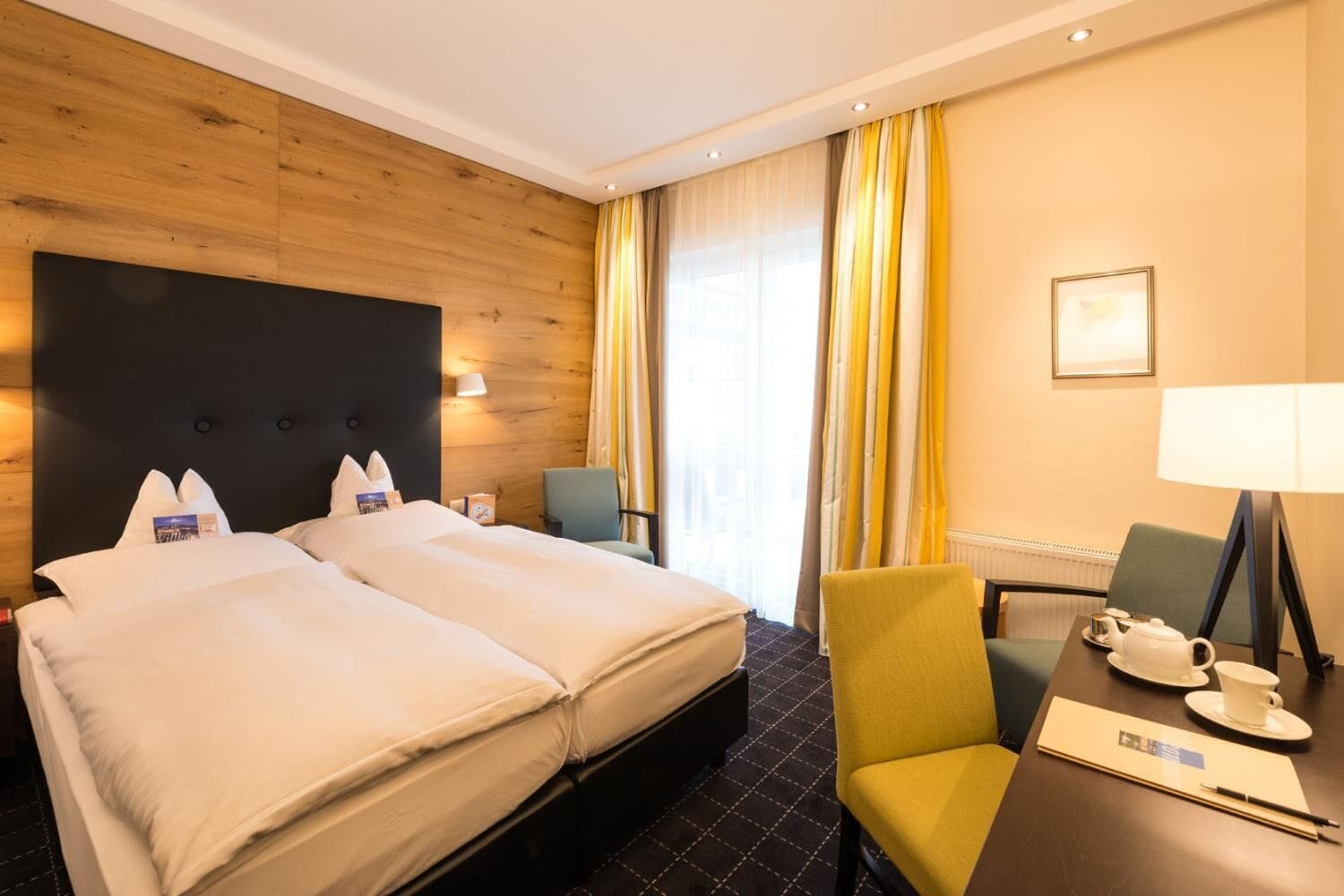 Ein Hotelzimmer mit Doppelbett, zwei Nachttischen, einem Schreibtisch mit Stuhl, einer Lampe und einem Vorhangfenster.