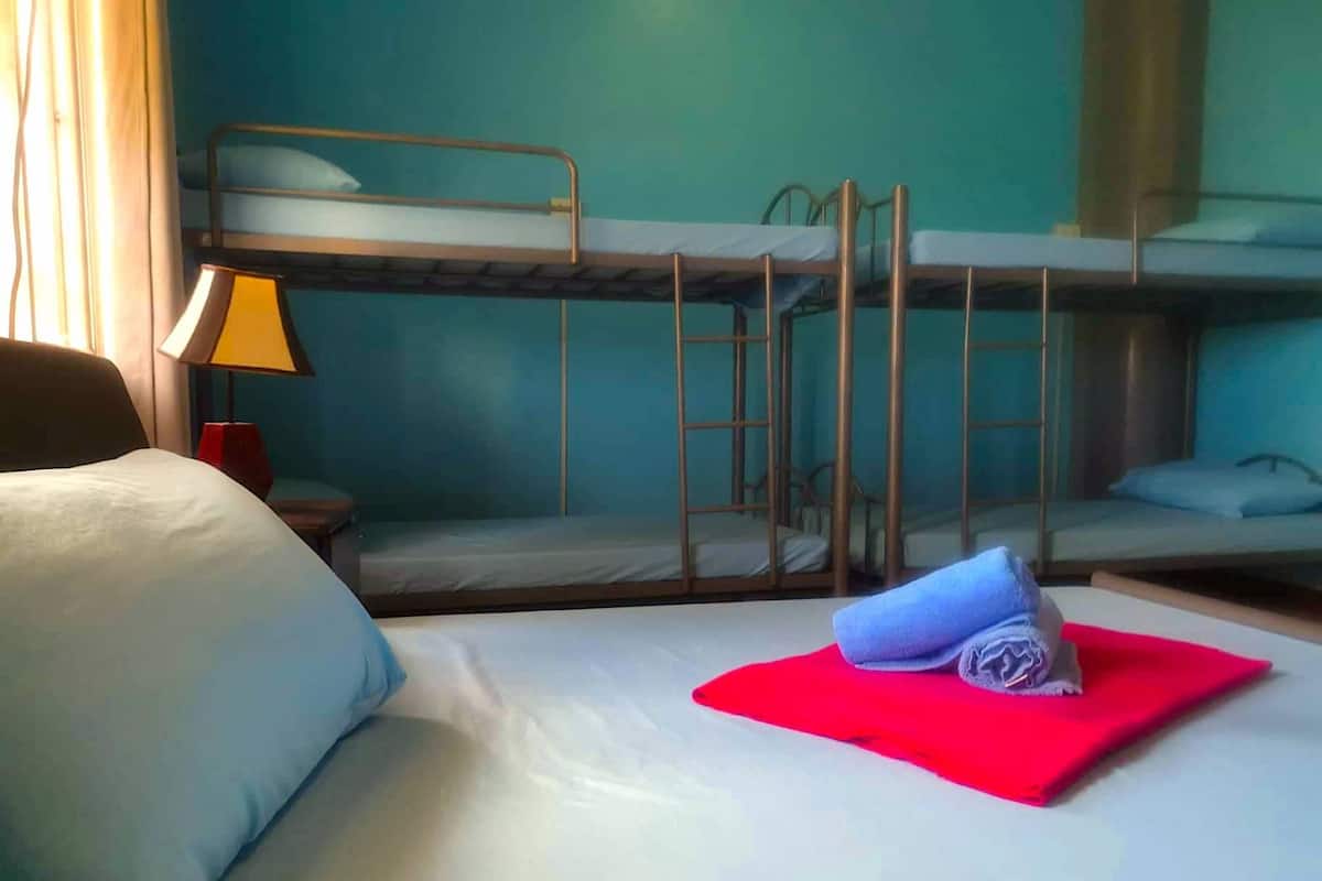 Une chambre d’hôtel avec un lit superposé, un bureau avec une lampe et un lit bien fait, agrémenté d’une serviette rouge.