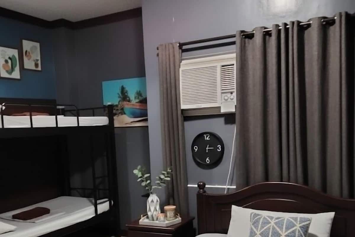 Une chambre d’hôtel avec un lit simple, un lit superposé plus petit, une table de chevet avec une horloge et une fenêtre avec des rideaux.