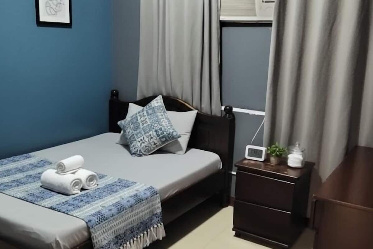 Une chambre à coucher avec un lit, une table de chevet avec une radio, une petite plante et une fenêtre avec des rideaux.