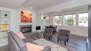 A modern living room with a fireplace, grey sofas, a dining table, and large windows offering a view of the outdoors. - Relaxing Lakefront Suite (Federal Way)