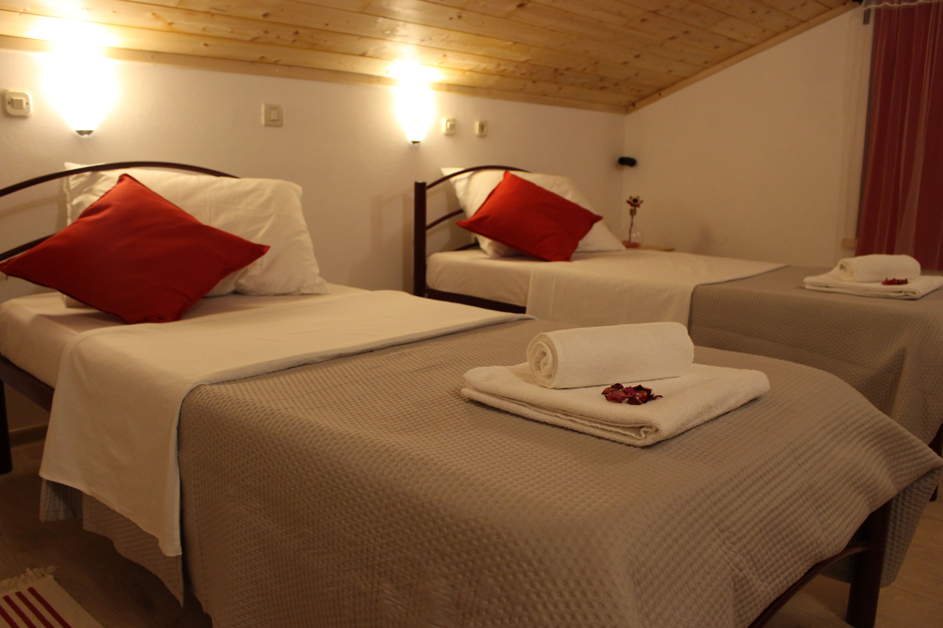 A hotel room with two single beds, each with a red pillow and a white blanket. There is a small table with a flower vase and a folded towel.