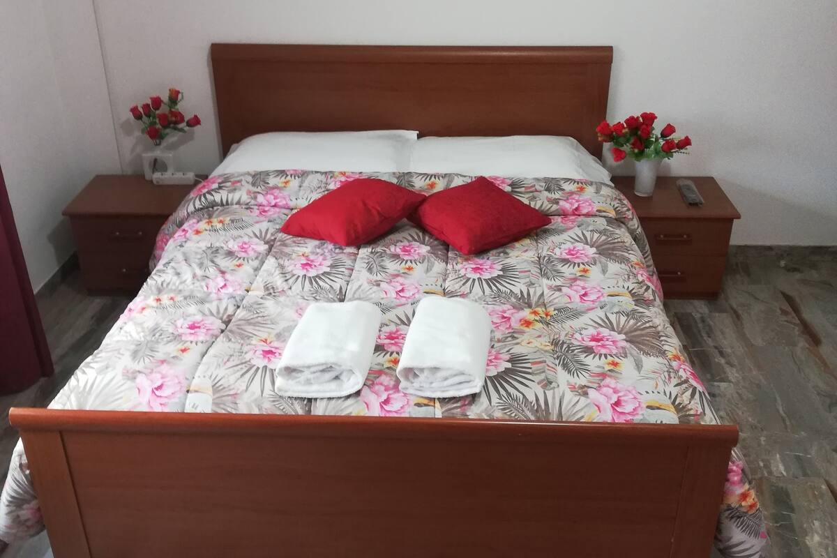A neatly made bed with floral bedding, two red pillows, and two towels. There are two bedside tables with vases of red flowers. A cityscape painting hangs above the bed.