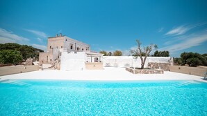Une piscine avec une eau d’un bleu limpide, un bâtiment blanc aux murs de pierre et un arbre au premier plan.