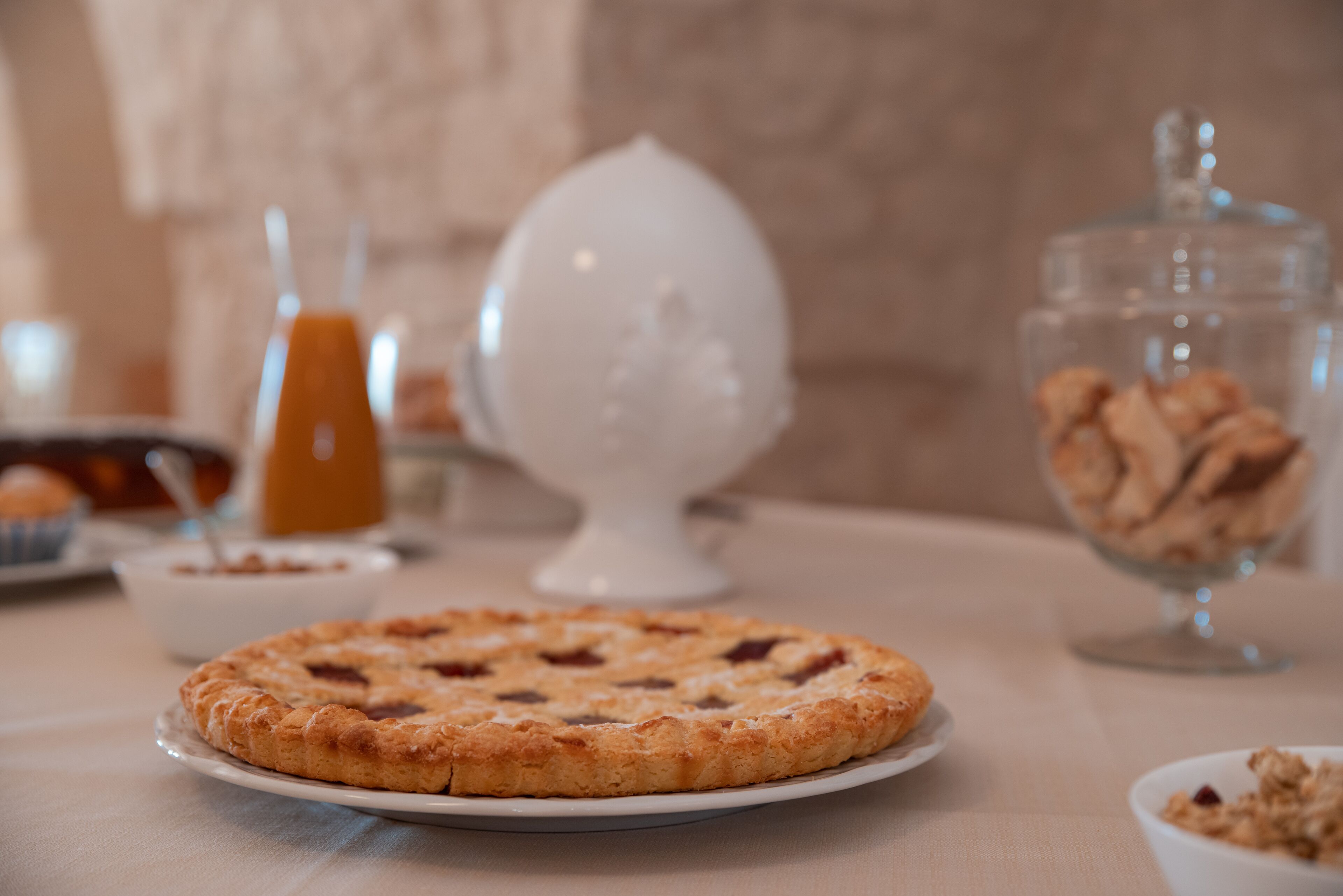 A breakfast setting with a round pastry on a plate, a glass container with cookies, and a glass of orange juice.