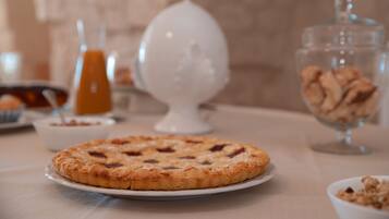 Une table mise pour le petit-déjeuner avec une pâtisserie ronde sur une assiette, un récipient en verre contenant des biscuits et un verre de jus d’orange.