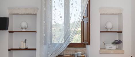 Una habitación con un escritorio de madera, una silla, un ventanal con cortinas traslúcidas y estantes con adornos.