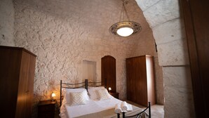 Une chambre avec un mur en pierre, un lit recouvert de draps blancs, une table de chevet avec une lampe et une armoire en bois.