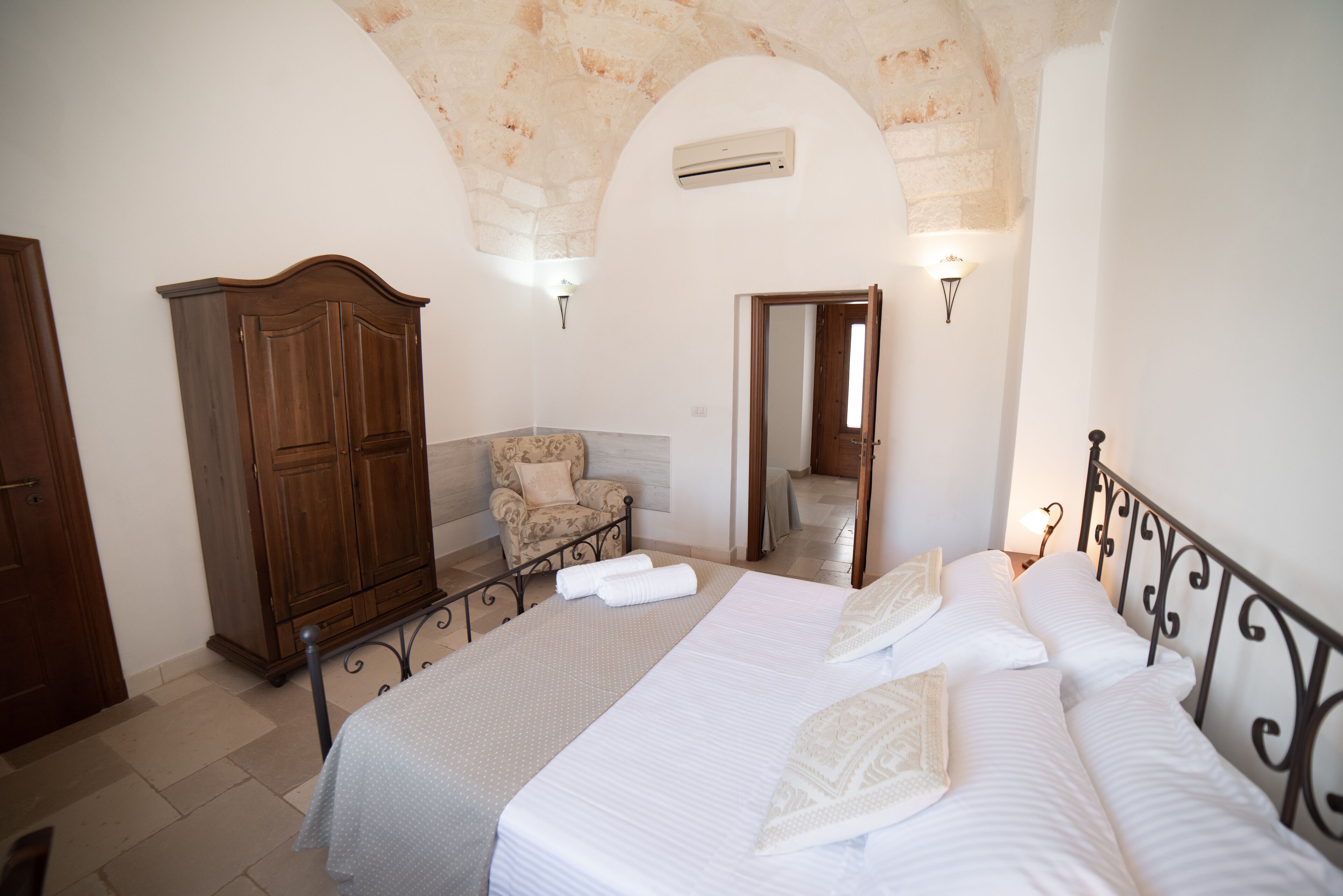 A bedroom with a large bed, a wooden wardrobe, a chair, and a door leading to another room.