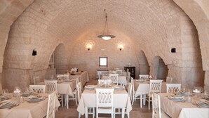 Un espace repas avec des tables et des chaises blanches, des murs en pierre et un lustre central.