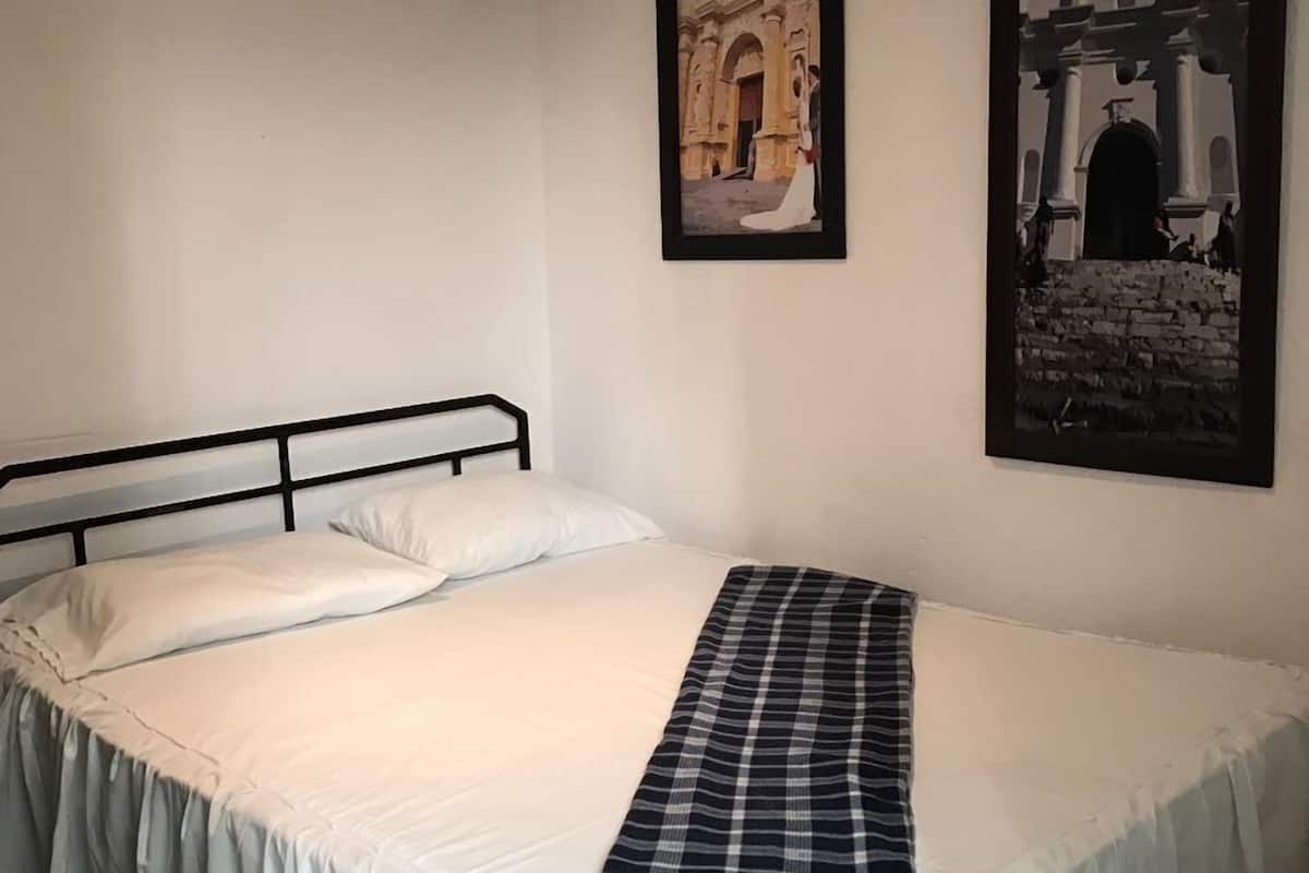 A single bed with white bedding and a plaid blanket, a black metal headboard, and two framed pictures of historical buildings on the wall.