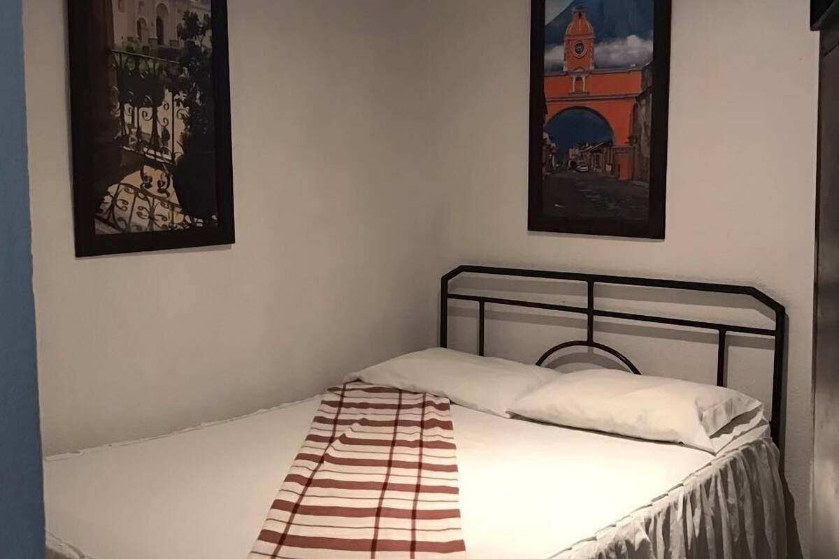 A single bed with a striped blanket, white pillows, and a black metal headboard in a room with white walls. There are two framed pictures on the walls, one depicting a building and the other a landscape with a tower.