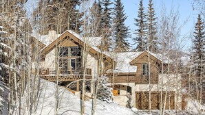 A large wooden cabin with multiple windows and a balcony, surrounded by snow-covered trees and a clear sky.