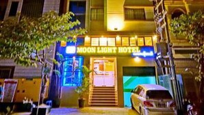 Un hôtel dont l’entrée est marquée par une enseigne bien visible indiquant « MOON LIGHT HOTEL », avec une voiture garée devant et un escalier menant à l’entrée.