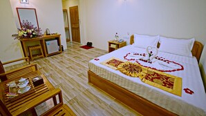 Une chambre d’hôtel avec un lit bien fait, une table de chevet en bois surmontée d’un vase de fleurs, un petit bureau avec une lampe et une chaise en bois.