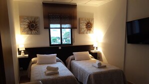A hotel room with two beds, a TV, and a window with a view of greenery. - Miramar Habitaciones (Aguilas)