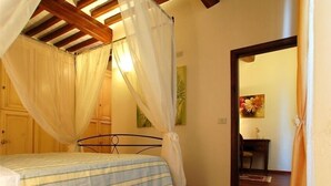 Un dormitorio con una cama bajo un dosel, vigas de madera en el techo, un escritorio con una silla y un cuadro en la pared.