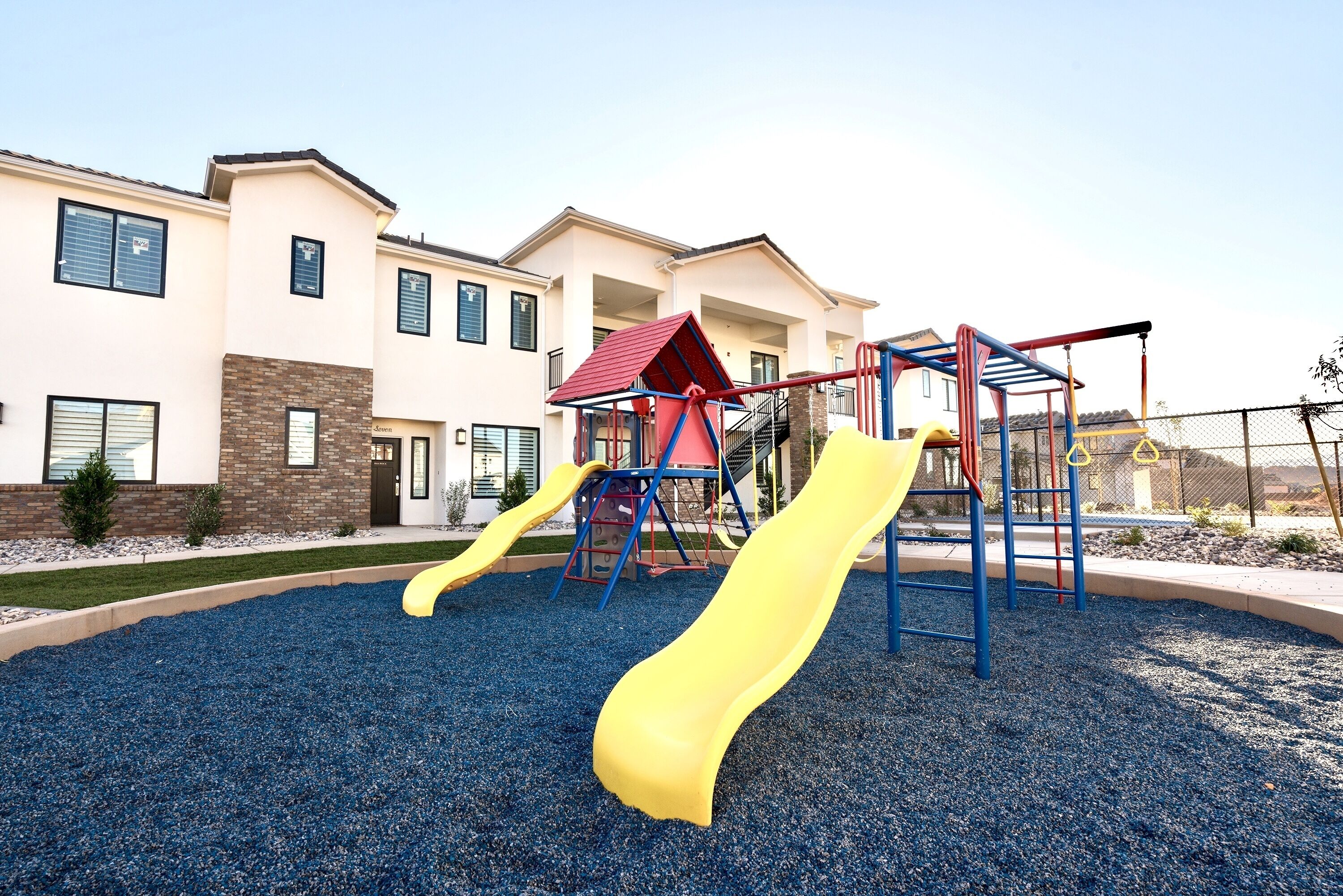 A playground with slides and climbing structures, surrounded by a building with multiple windows and a brick facade.
