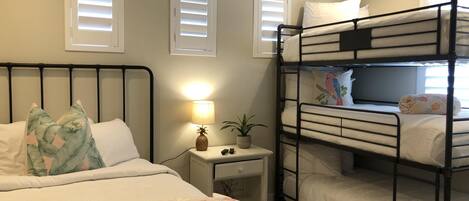 A bunk bed room with a single bed on the left and a bunk bed on the right. There is a nightstand with a lamp and a small plant.