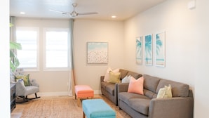 A living room with a grey rocking chair, a brown sofa, and a blue ottoman. There is a ceiling fan, a window with white curtains, and a wall-mounted picture. - ❀Aloha❀ - Tropical Island Retreat | POOL IS OPEN!!! Great Family Friendly Vacation! (Washington)