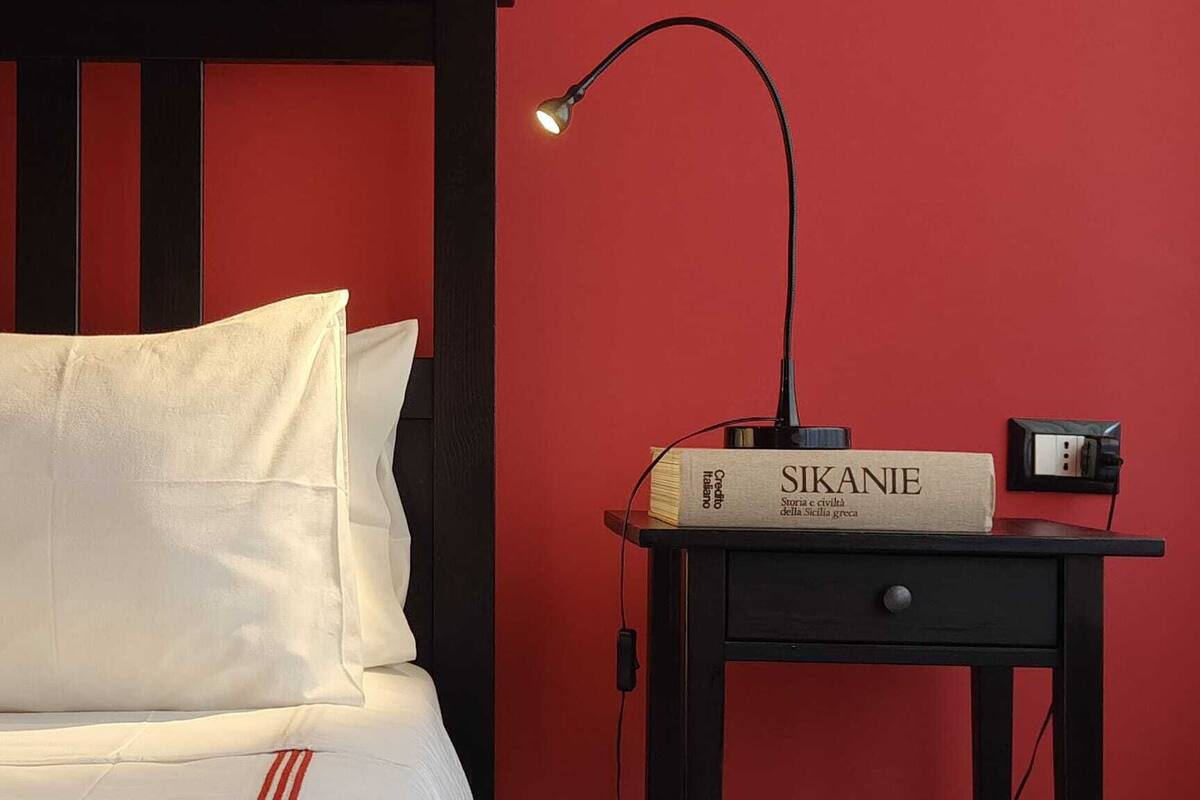 Un lit avec une literie blanche, une table de chevet avec une lampe et un livre intitulé « SIKANIE » posé sur la table.