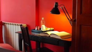 Une pièce avec un mur rouge, un radiateur, une chaise en bois, un bureau avec un livre, une lampe et une bouteille.