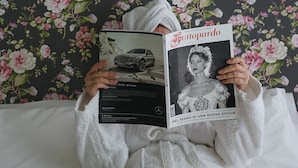Une personne allongée sur un lit, devant un mur recouvert d’un papier peint à motifs floraux, et qui lit un magazine.