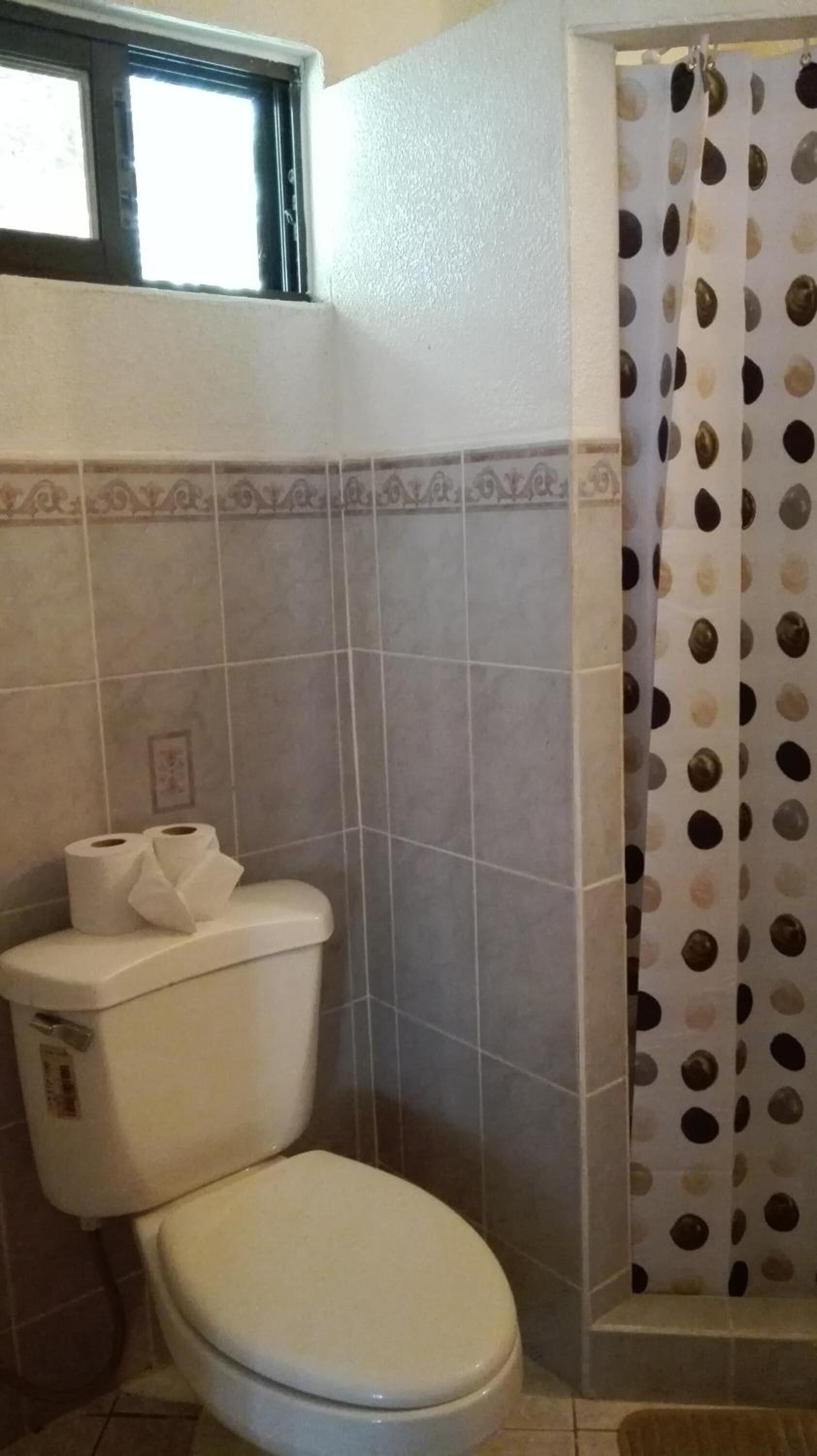 Un baño con inodoro, cortina de ducha estampada con lunares y una ventana sobre el inodoro.