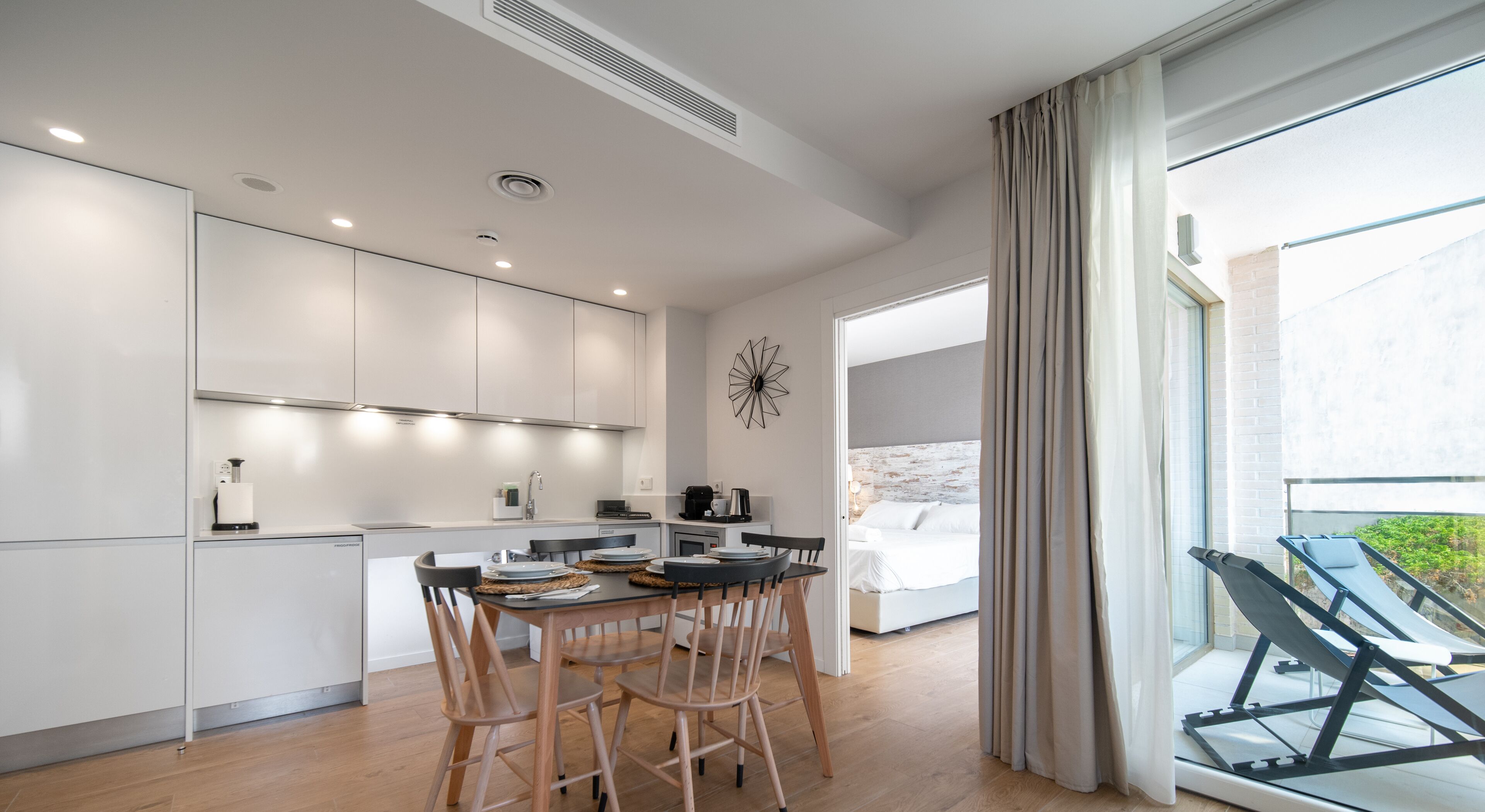Una cocina moderna con armarios blancos, un comedor con sillas de madera y un amplio ventanal con cortinas.