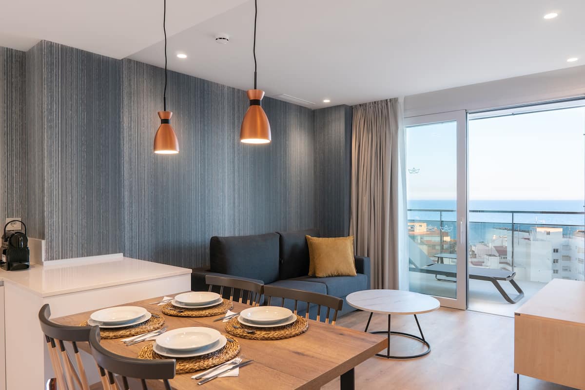 A modern dining area with a wooden table set for four, a sofa with cushions, and a balcony with a view.