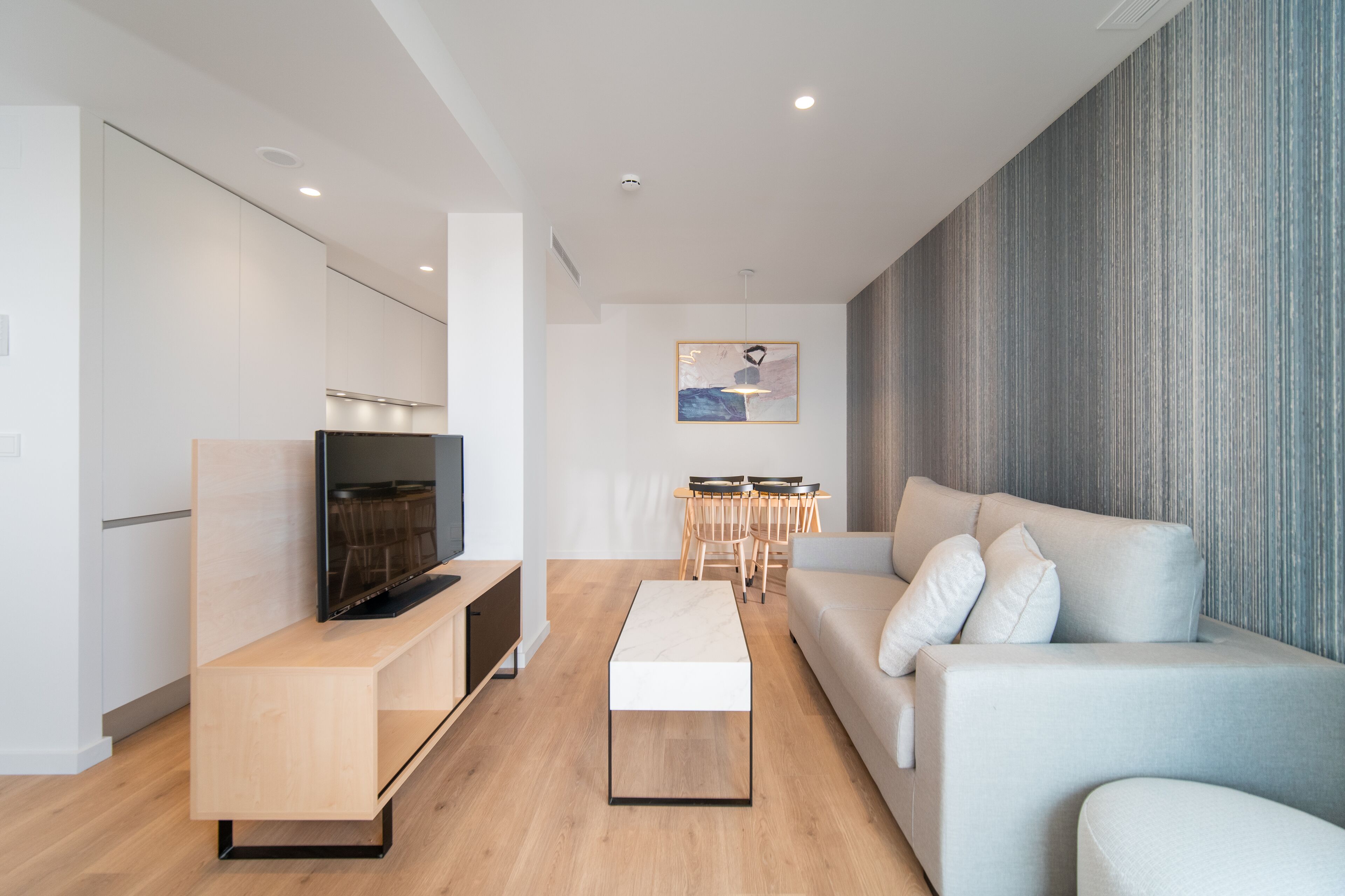 A modern living room with a flat-screen TV, a light-colored sofa, a coffee table, and a dining area with a wooden table and chairs.