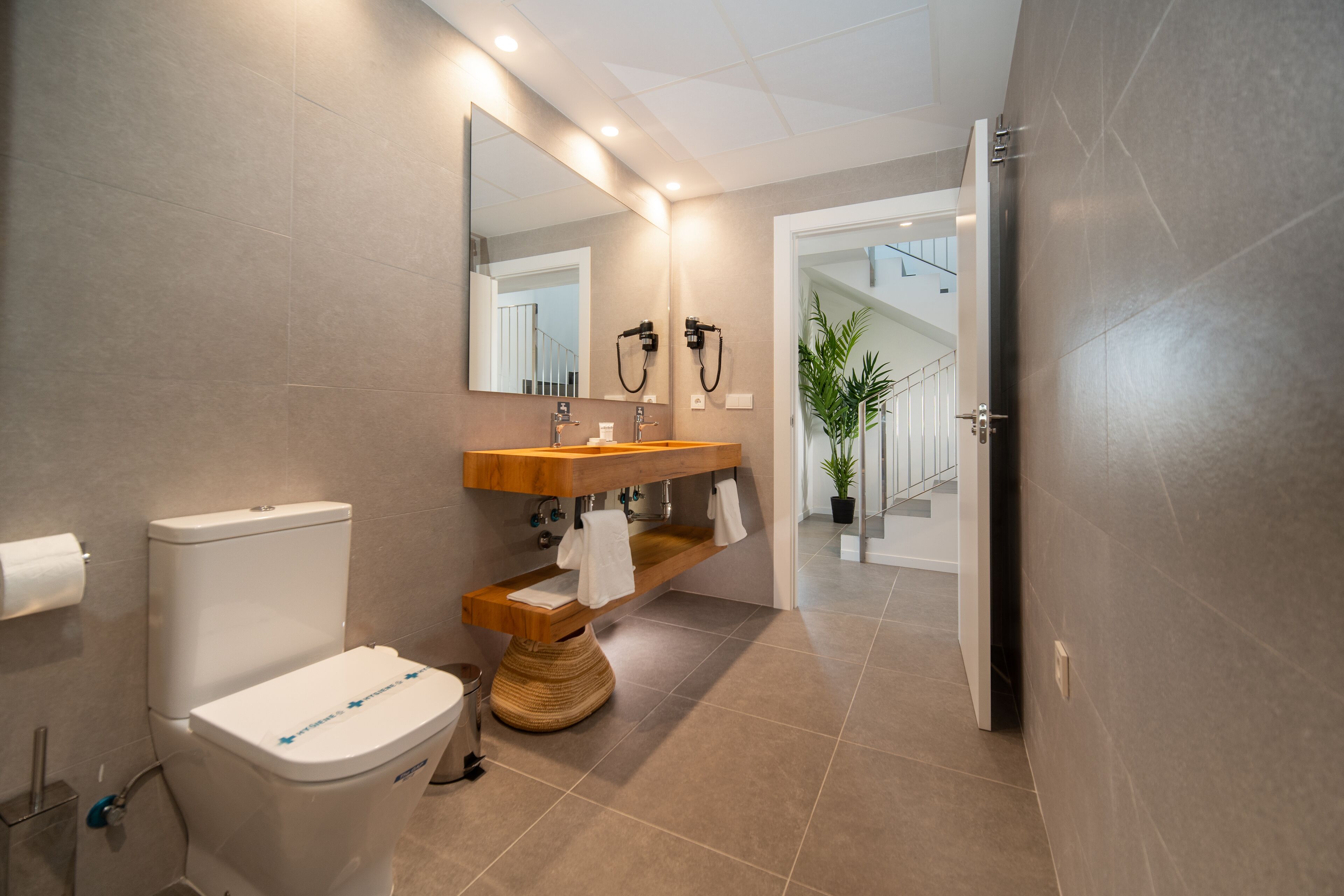 Un bagno moderno con WC, lavandino e specchio. Si nota una pianta sullo sfondo e una scala che conduce a un altro piano.