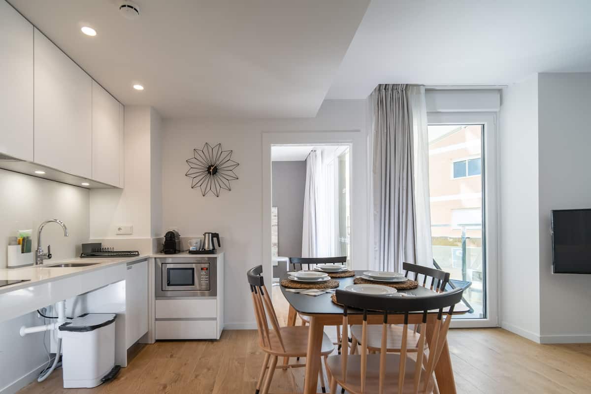 A modern kitchen with a built-in oven, microwave, and sink. There is a dining area with a table and chairs, and a TV mounted on the wall.