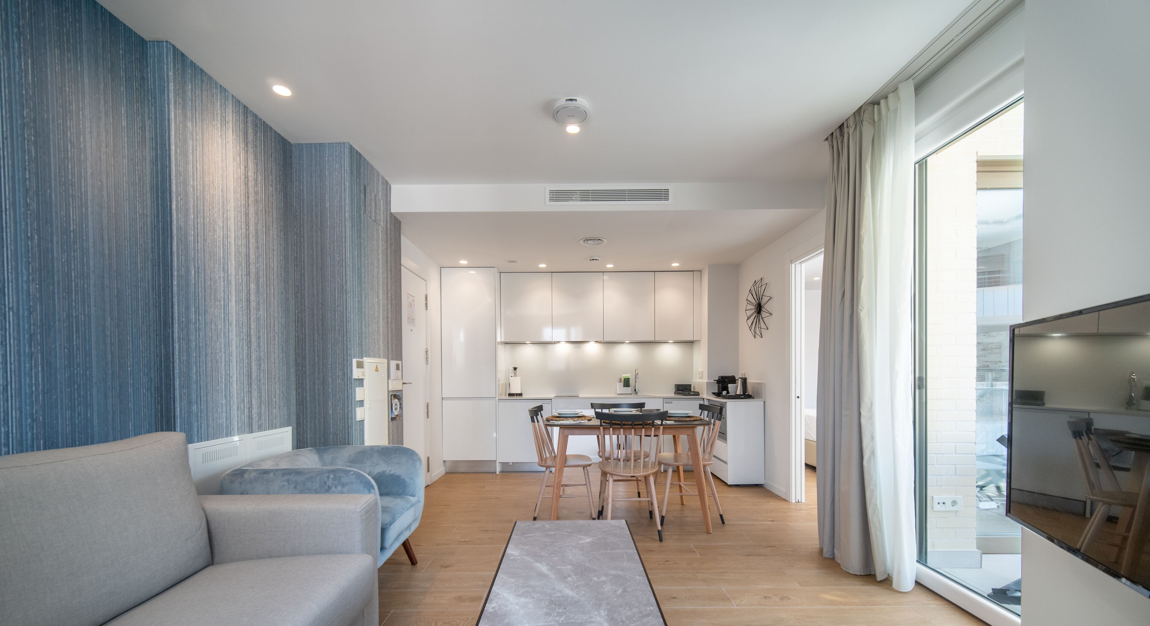 Una sala de estar moderna con un sofá, un comedor con mesa y sillas, una cocina con armarios blancos y un gran ventanal con cortinas.