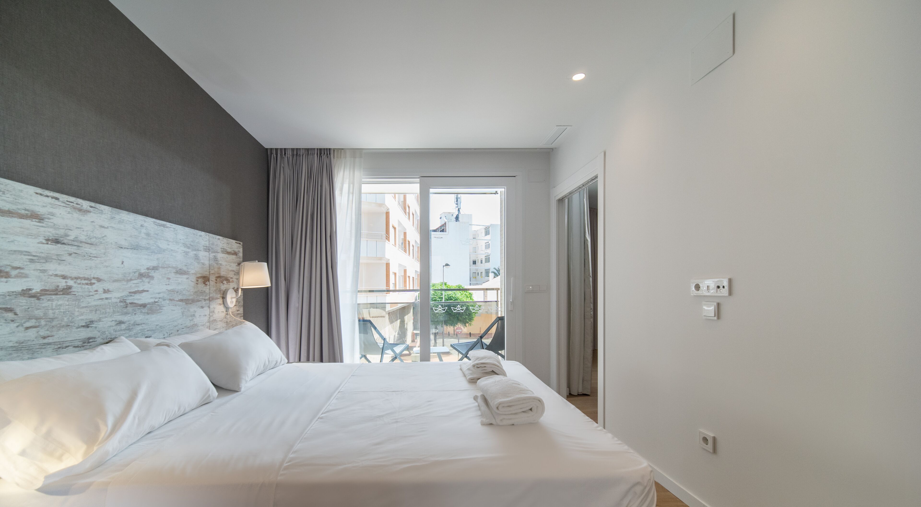 Une chambre d’hôtel avec un grand lit, une tête de lit au style rustique, un balcon avec des chaises et une vue sur les bâtiments environnants.