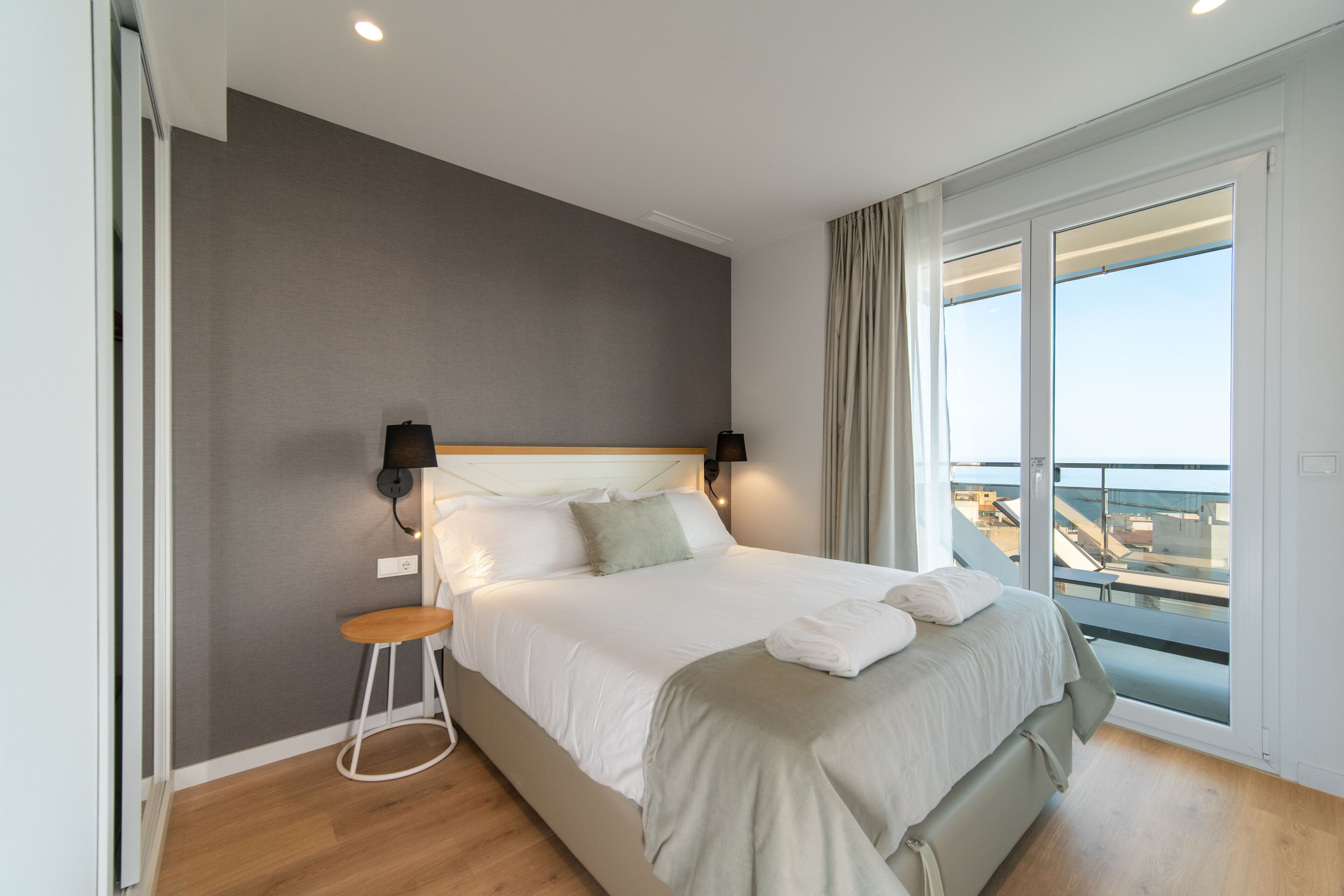 A modern hotel room with a large bed, a small stool, and a view of the sea through a sliding glass door.