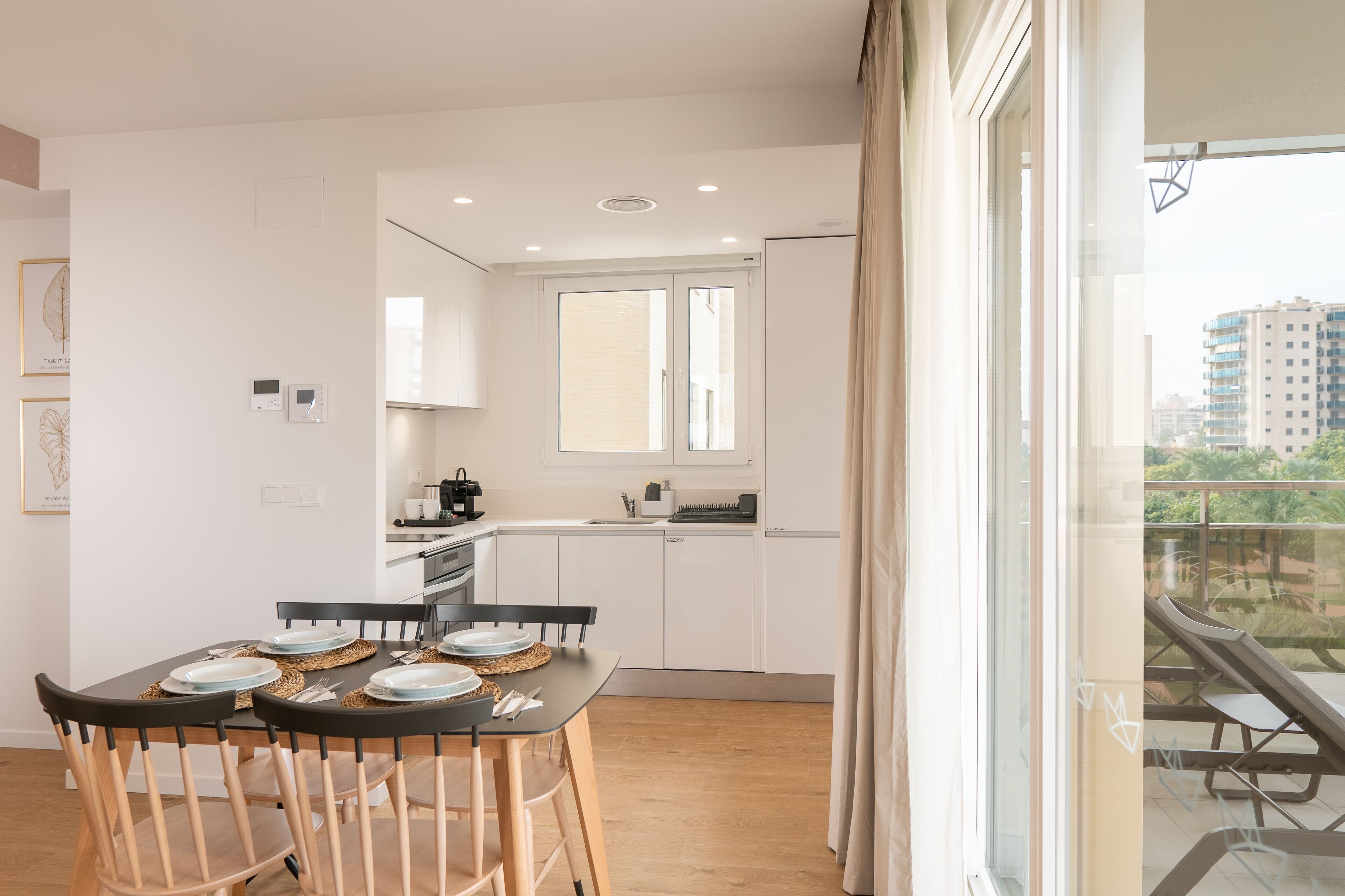 Une cuisine moderne avec des meubles blancs, une table à manger dressée et une vue sur un balcon aménagé avec du mobilier d’extérieur.