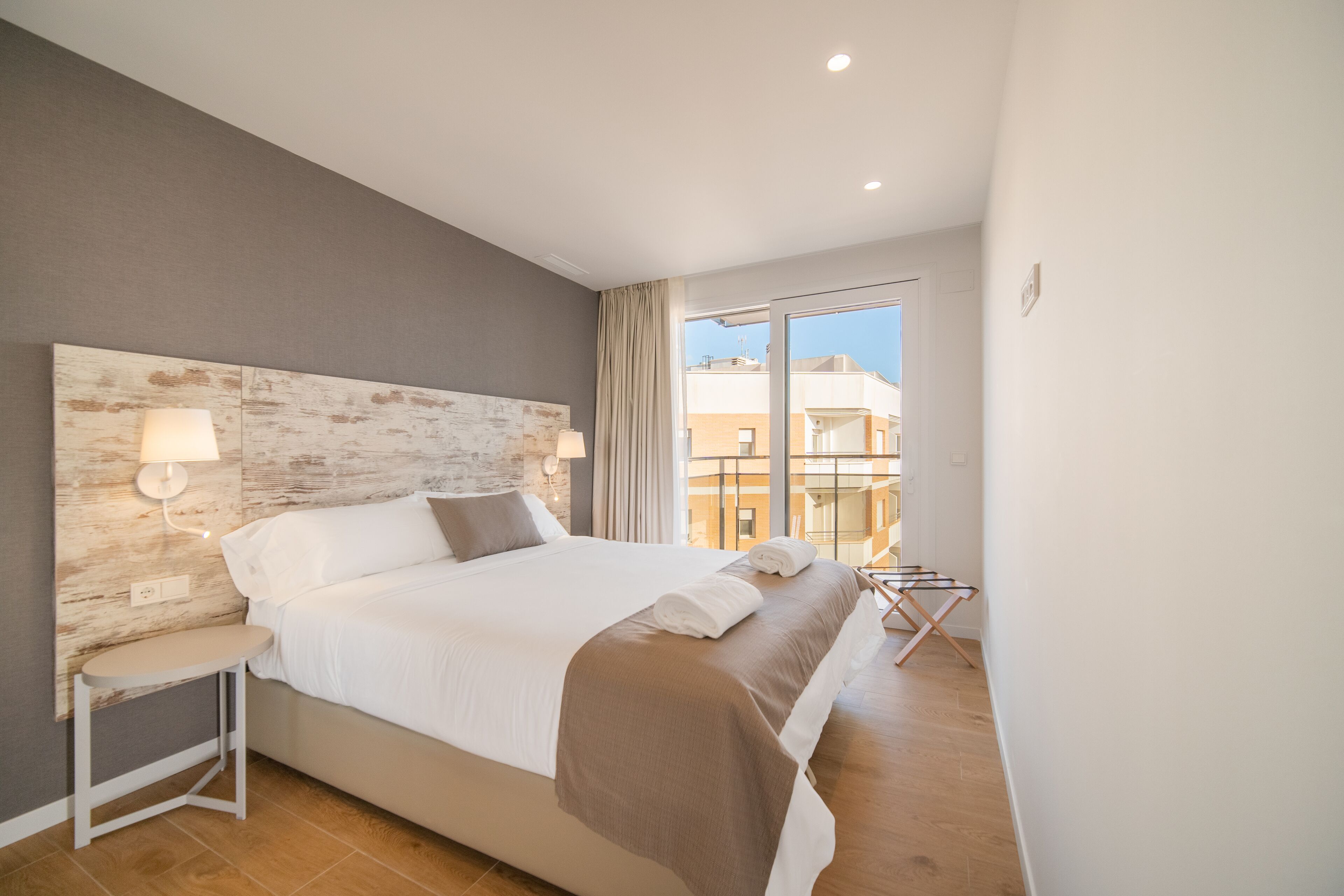 Ein modernes Hotelzimmer mit einem großen Bett, einem Nachttisch, einer Lampe und Blick auf die Stadt durch große Fenster.