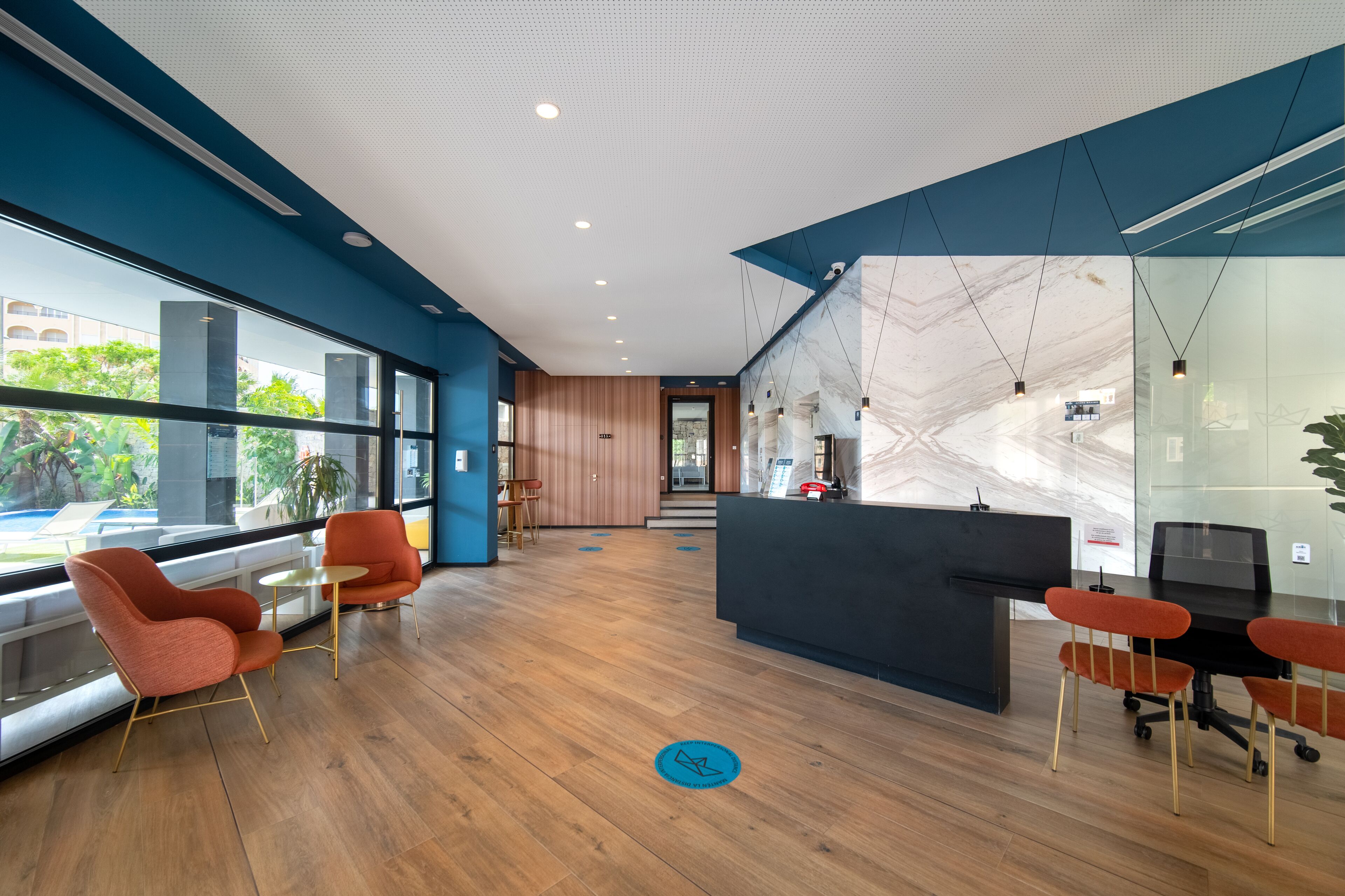 A modern reception area with a wooden floor, orange chairs, a black reception desk, and large windows offering a view of greenery outside.