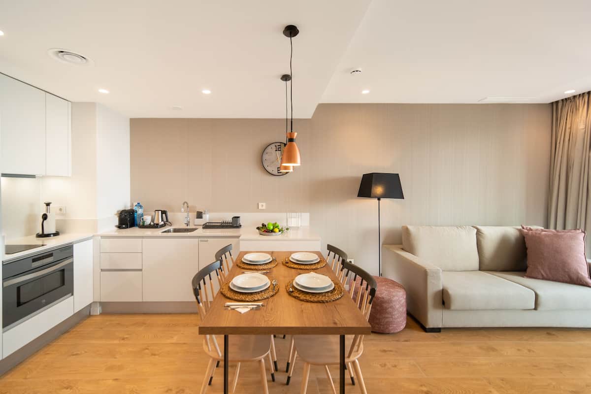 A modern kitchen with white cabinets, a wooden dining table set for four, and a sofa in the living area.