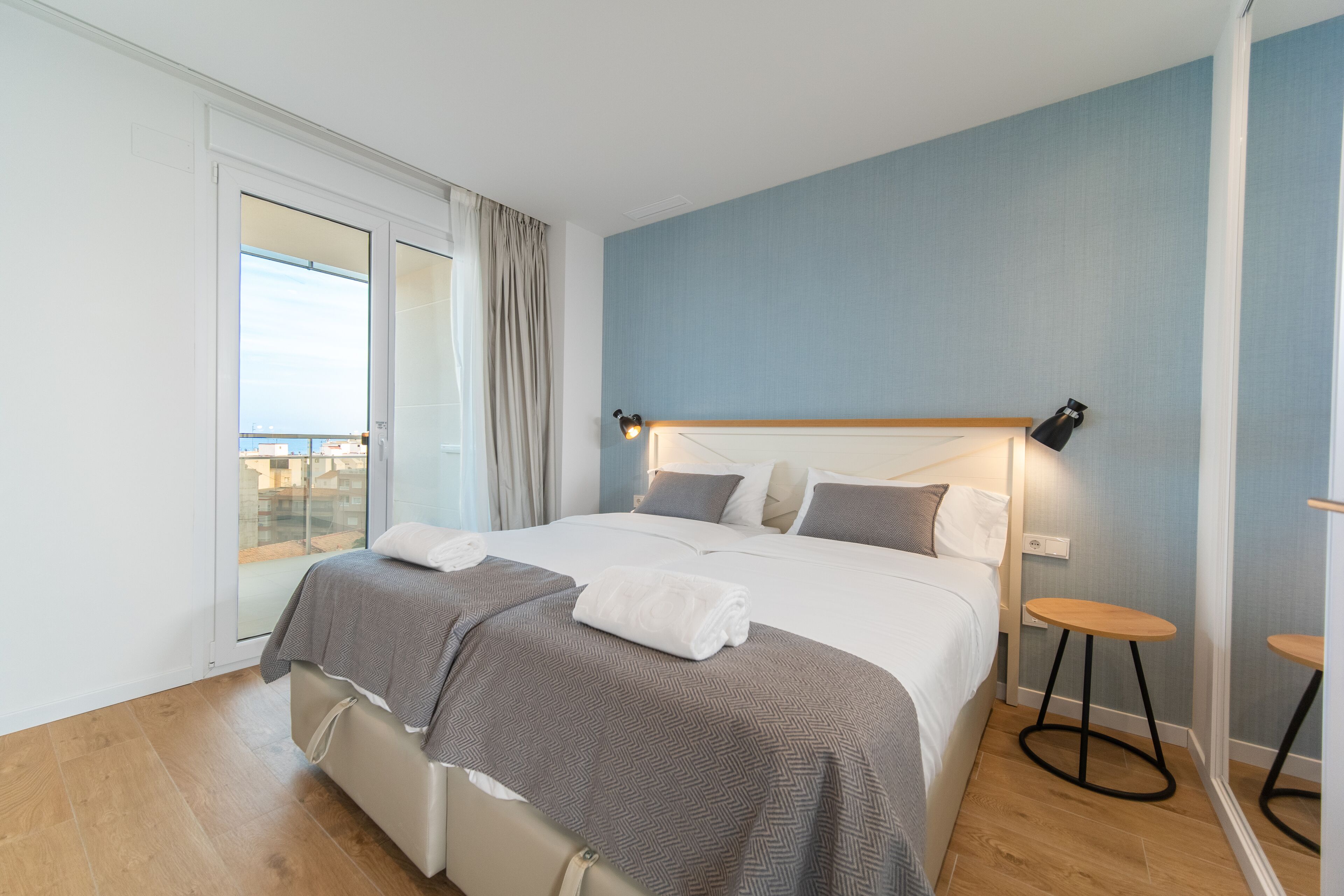 A modern bedroom with a large bed, a small stool, and a view through a sliding door.