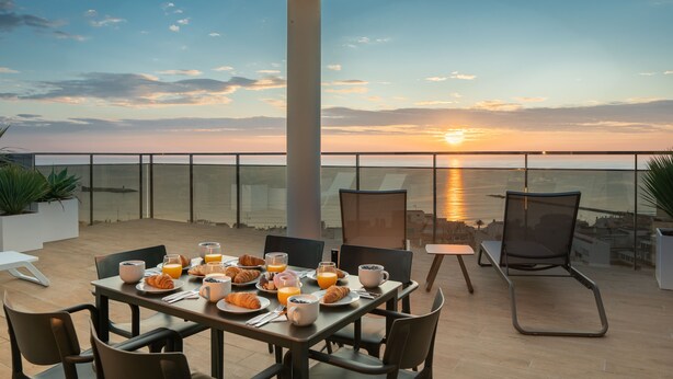 A breakfast table set with croissants, juice, and coffee, overlooking a sunset over the sea. - Ahoy Apartments (El Campello)