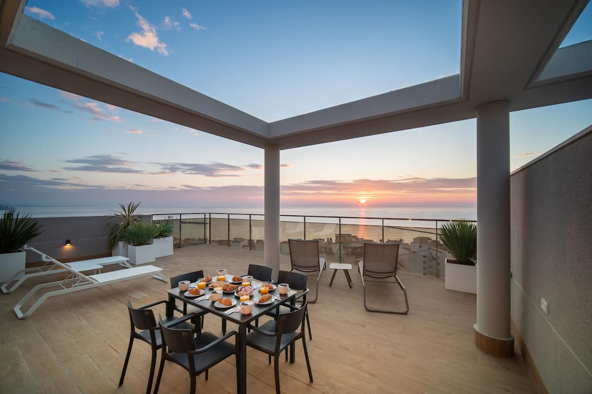 A rooftop terrace with a table set for breakfast, overlooking a sunset over the sea.