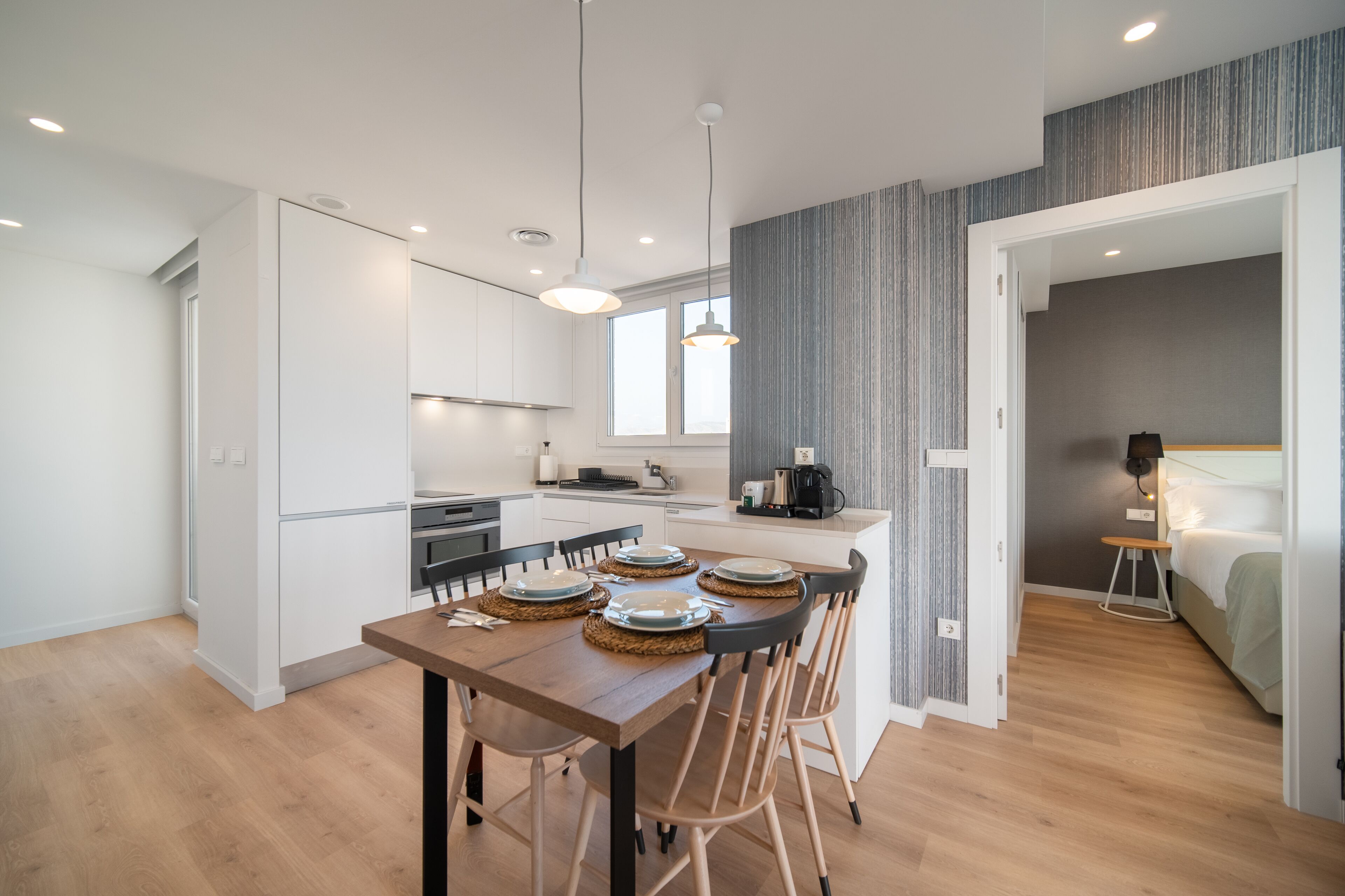 Una cocina moderna con armarios blancos, un comedor con mesa y sillas de madera, y un dormitorio con cama y mesitas de noche.
