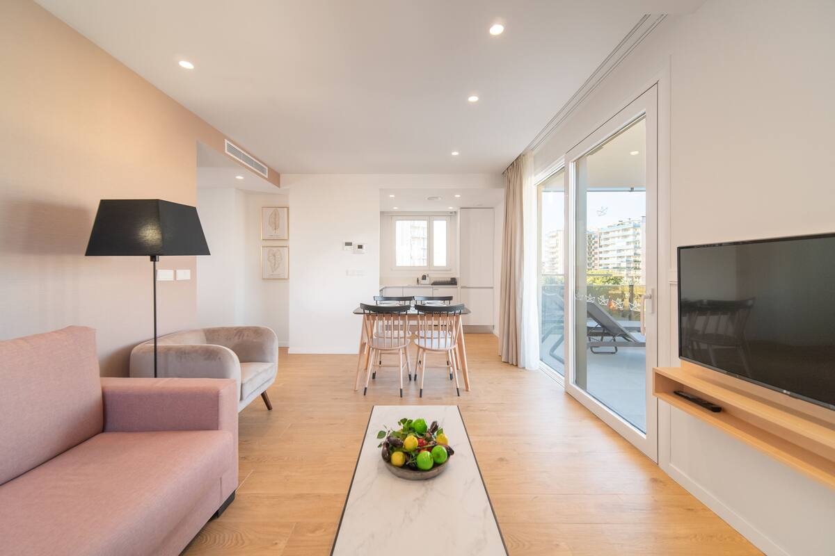A modern living room with a sofa, a coffee table with fruit, a TV, and a dining area with chairs and a table.
