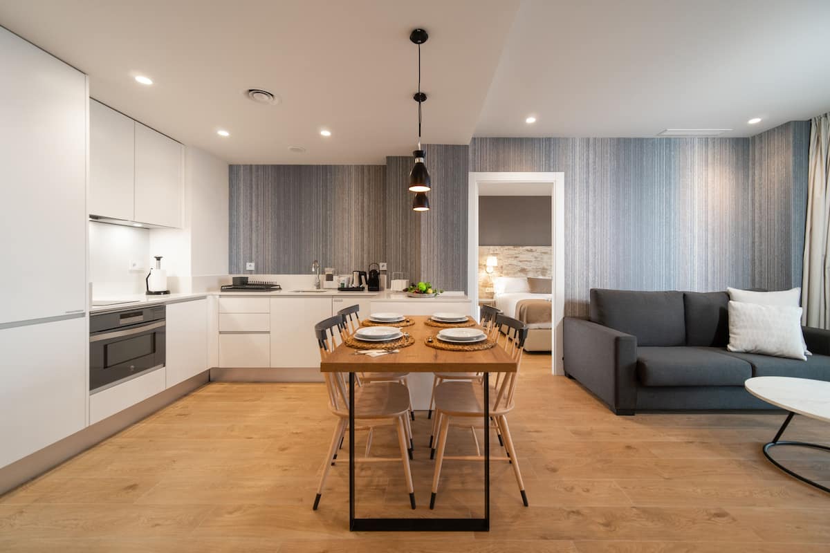 A modern kitchen with white cabinets, a wooden dining table set for four, and a grey sofa in the living area.