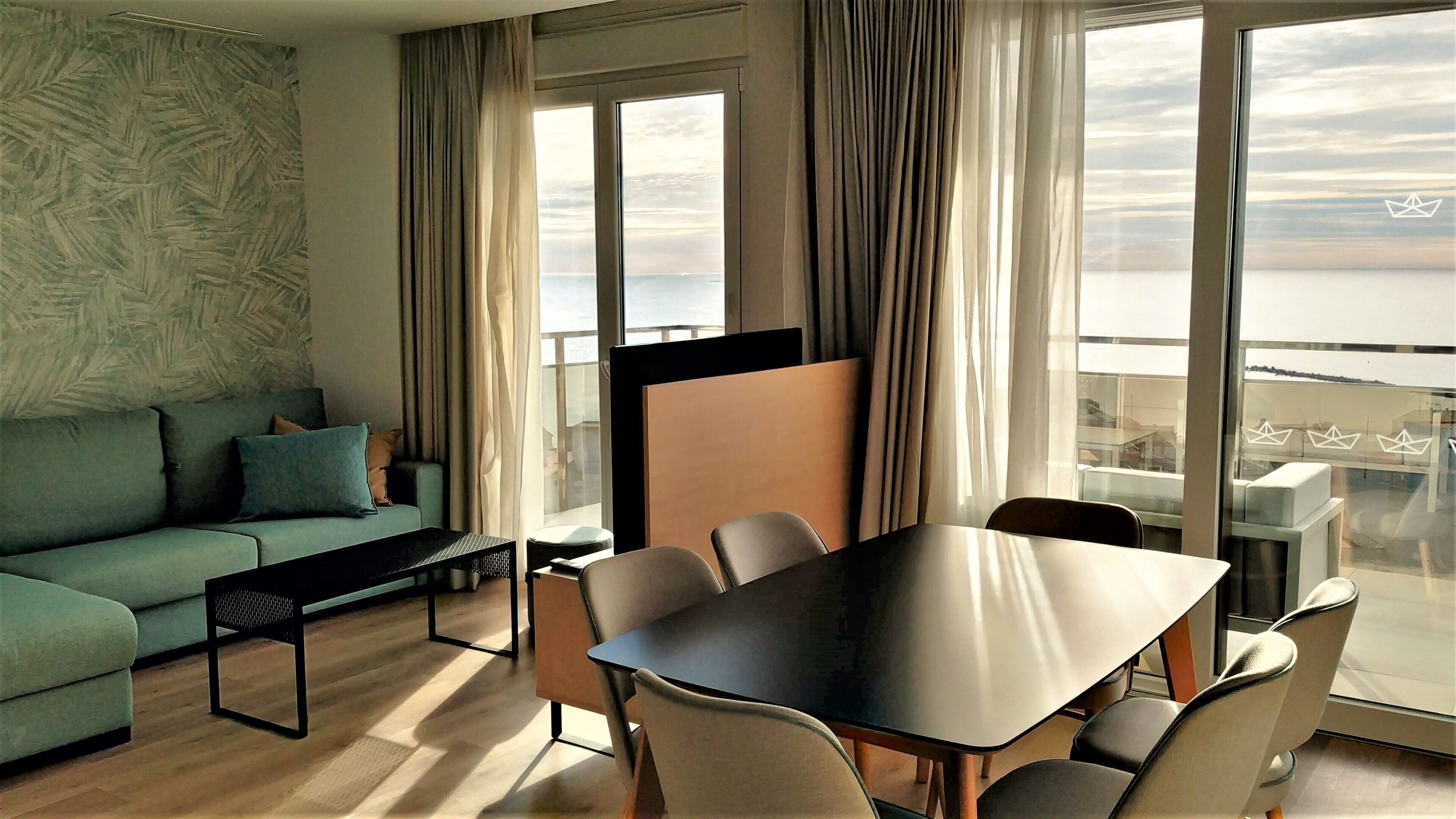 A modern living room with a sofa, a dining table with chairs, and large windows offering a view of the sea.