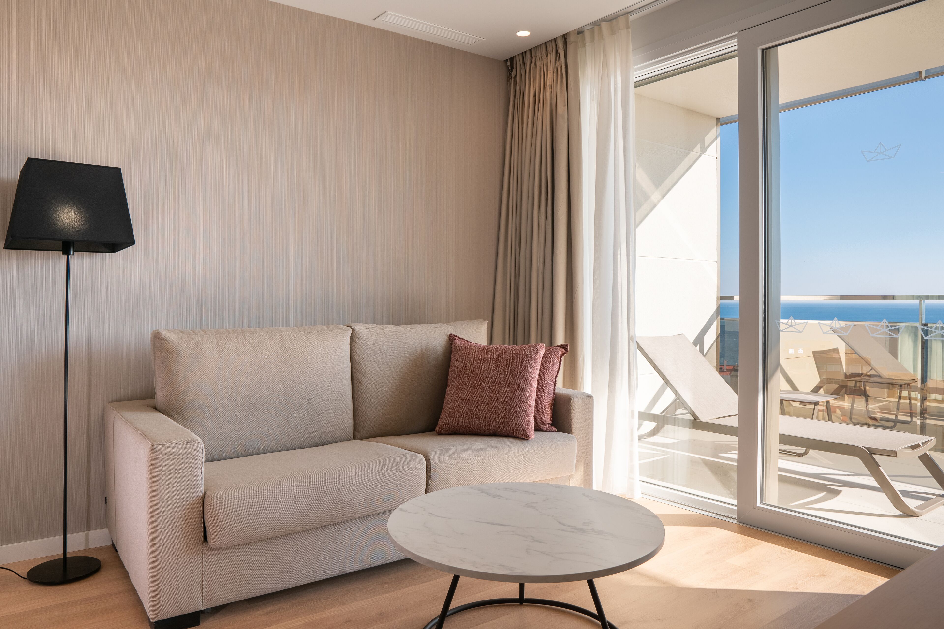 Een moderne hotelkamer met een beige bank, een ronde marmeren tafel en een groot raam met uitzicht op zee.
