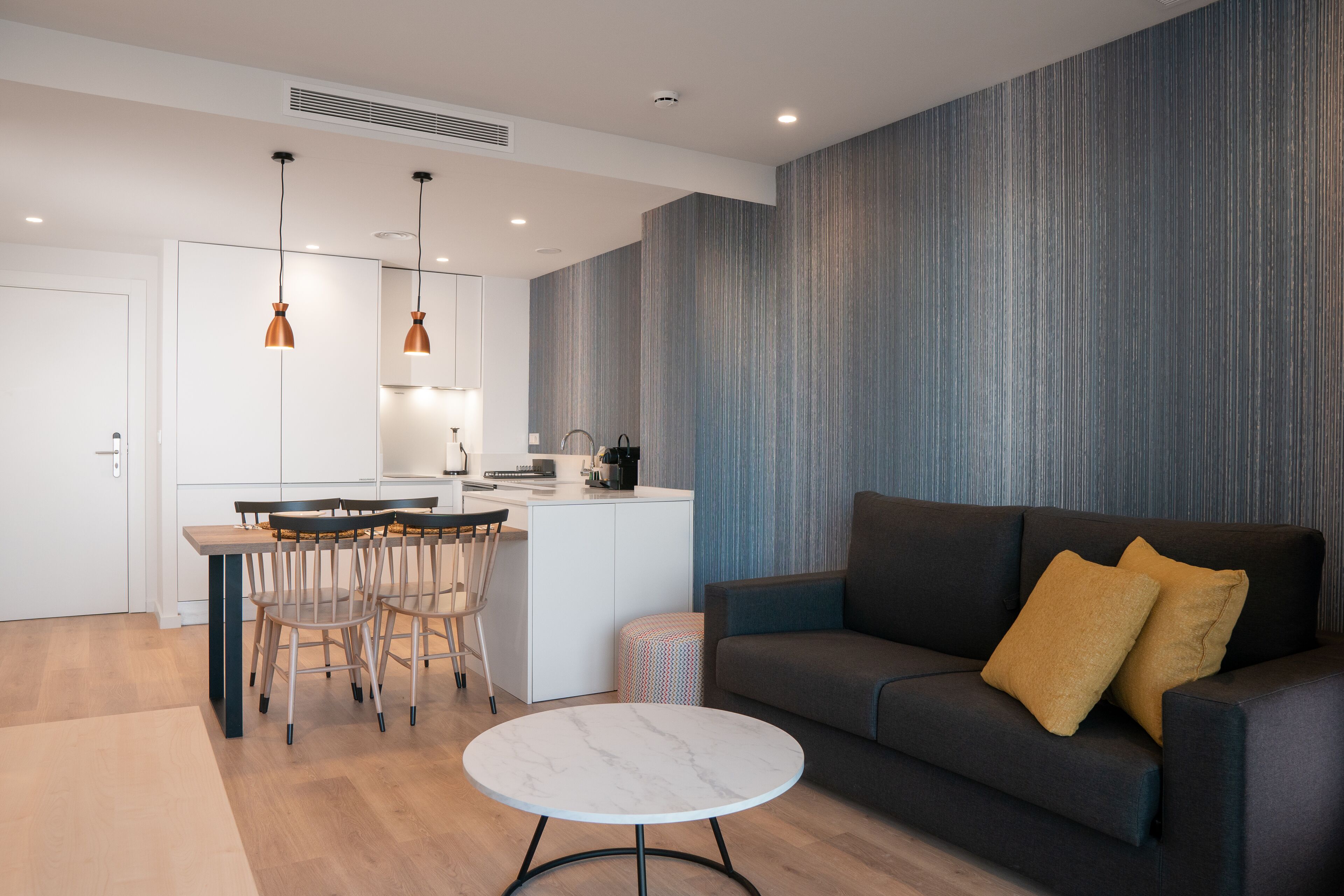 A modern living room with a dark sofa, a round coffee table, and a kitchen area with a dining set.